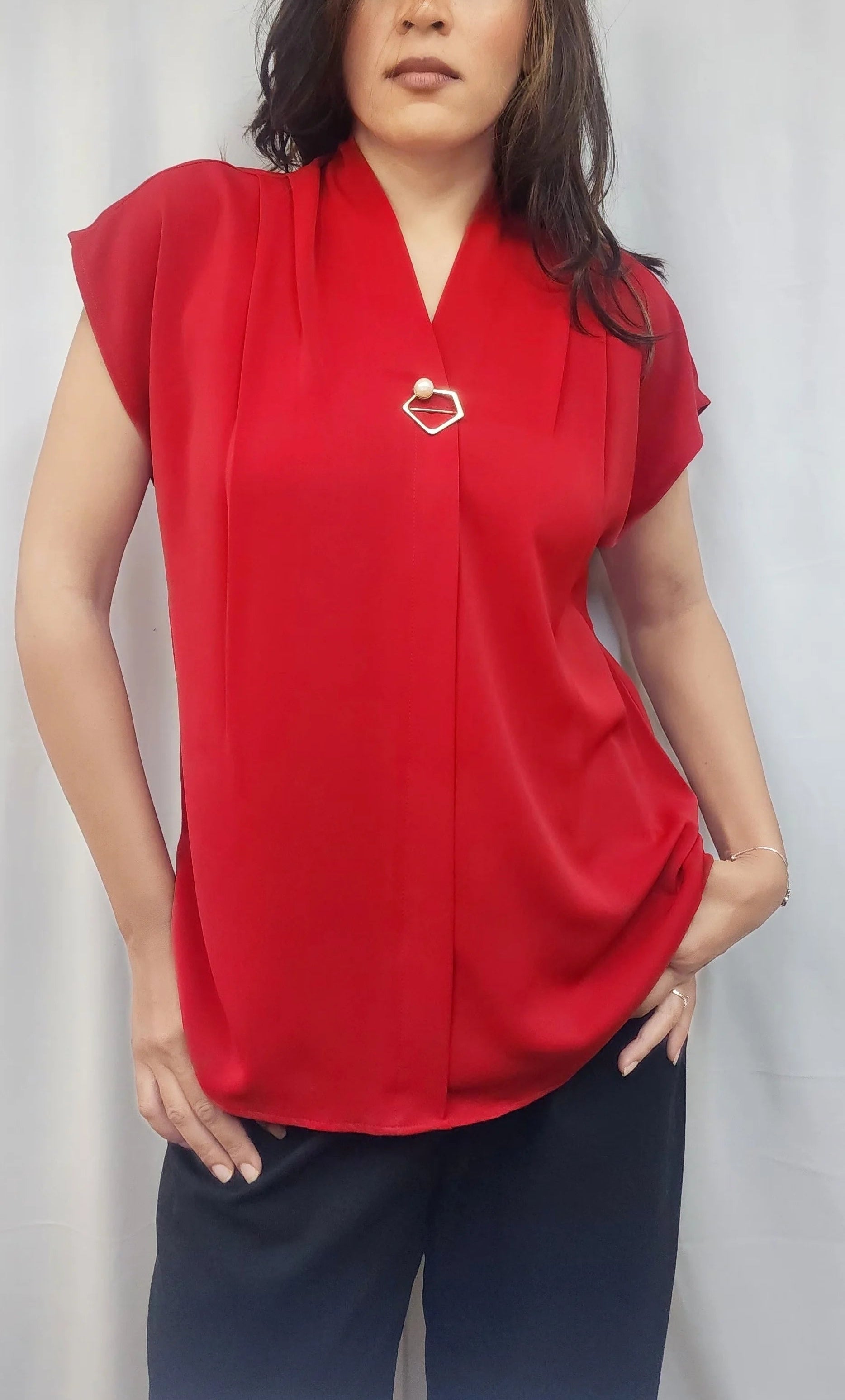 A woman wearing a bright red short-sleeve blouse with a V-neckline and a small gold heart-shaped brooch at the center chest, paired with dark navy pants, standing against a light gray curtain backdrop.