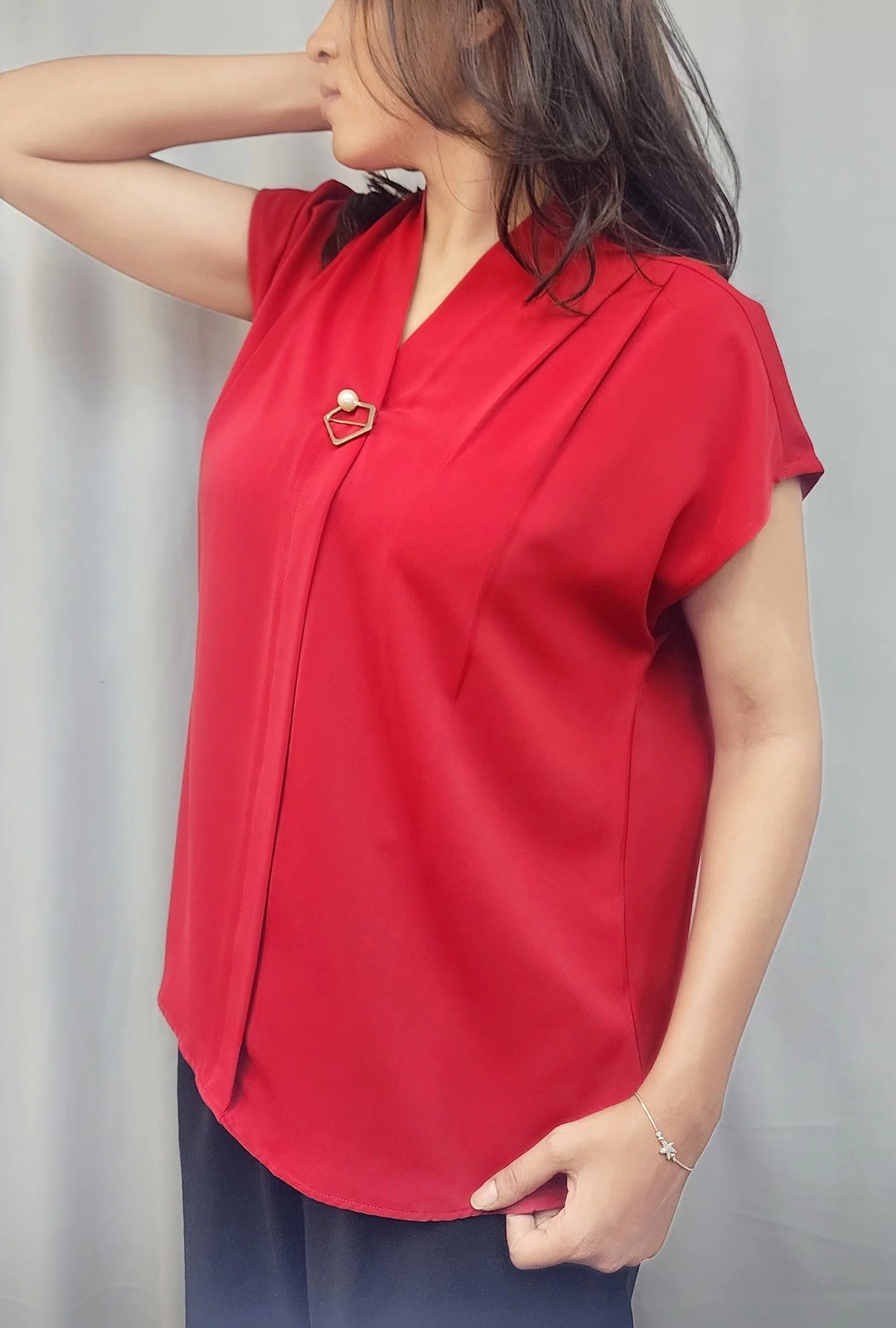 A woman wearing a bright red short-sleeve blouse with a V-neckline and a small decorative brooch at the collar, standing against a light gray curtain backdrop.