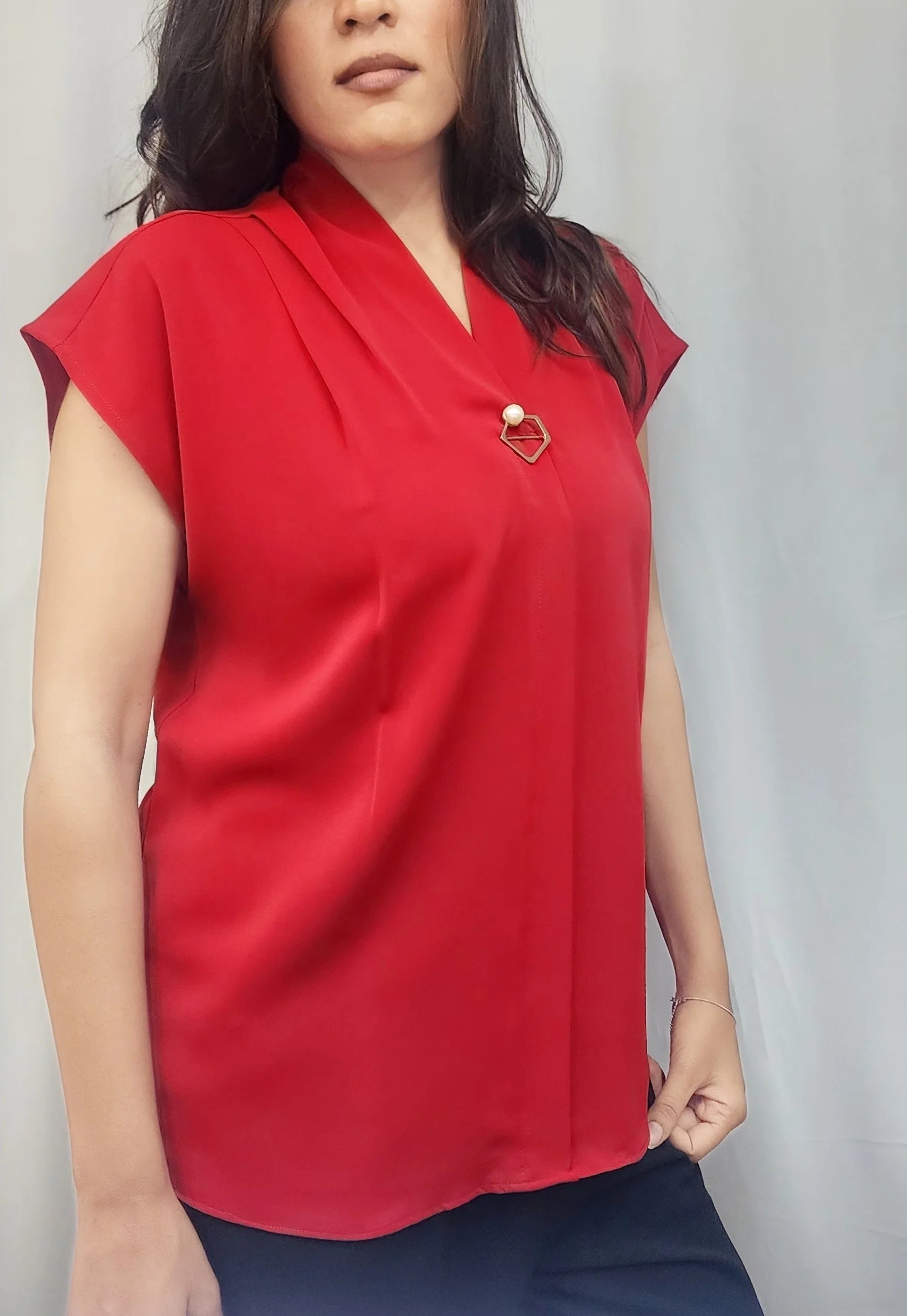 A woman wearing a bright red short-sleeve blouse with a V-neckline and a small gold heart-shaped brooch, paired with dark blue pants, standing against a light gray curtain backdrop.