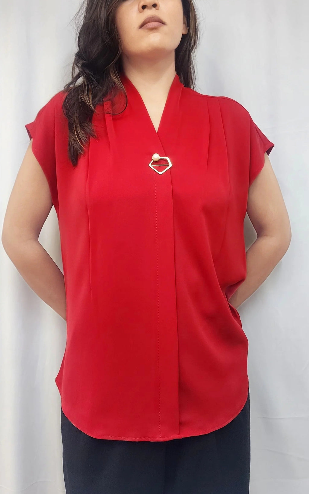 A woman wearing a bright red short-sleeve blouse with a V-neckline and a small metallic heart-shaped brooch on the chest, paired with dark pants, standing in front of a white curtain.