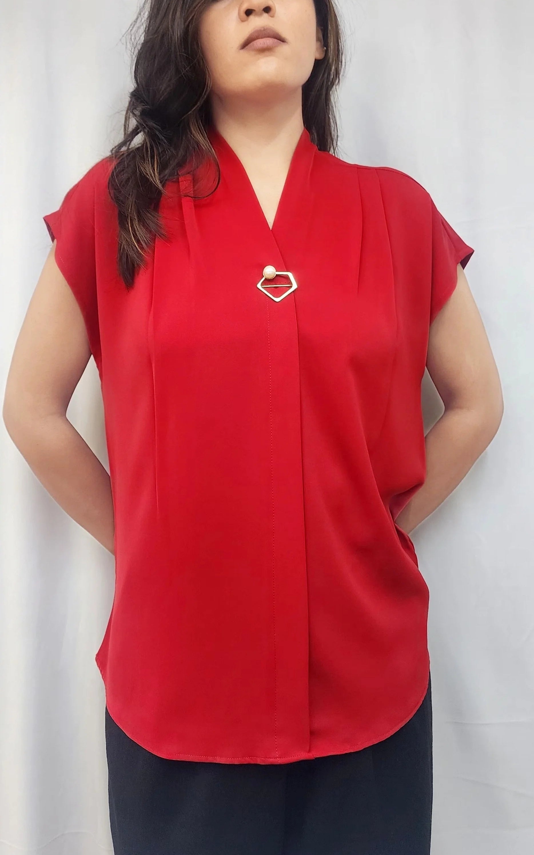 A woman wearing a bright red short-sleeve blouse with a V-neckline and a small metallic heart-shaped brooch on the chest, paired with dark pants, standing in front of a white curtain.