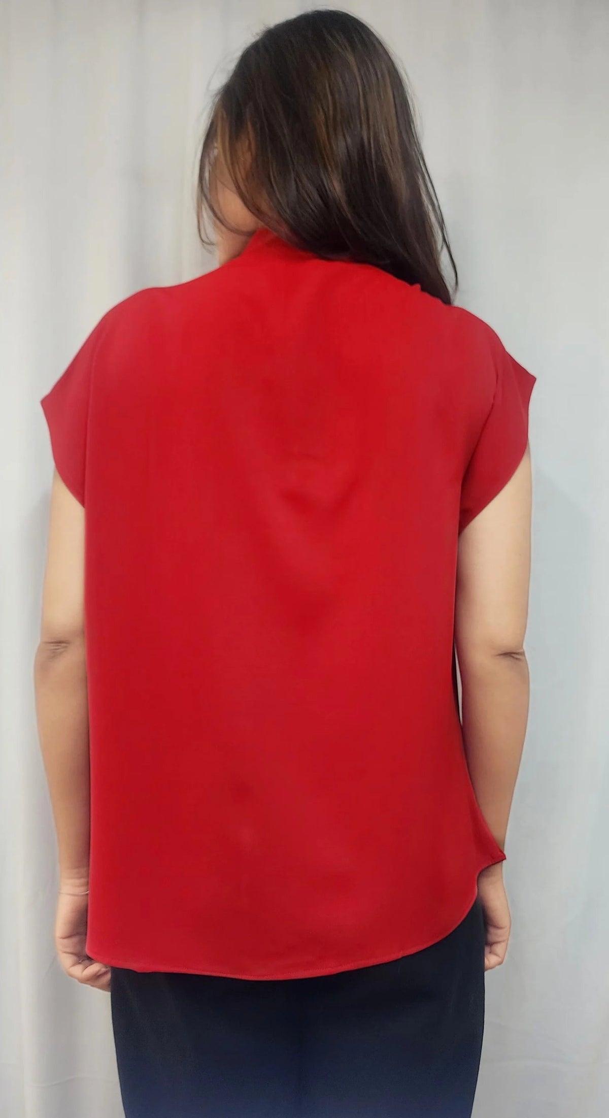 A woman viewed from behind wearing a bright red short-sleeve blouse and dark pants, standing against a light gray curtain background.