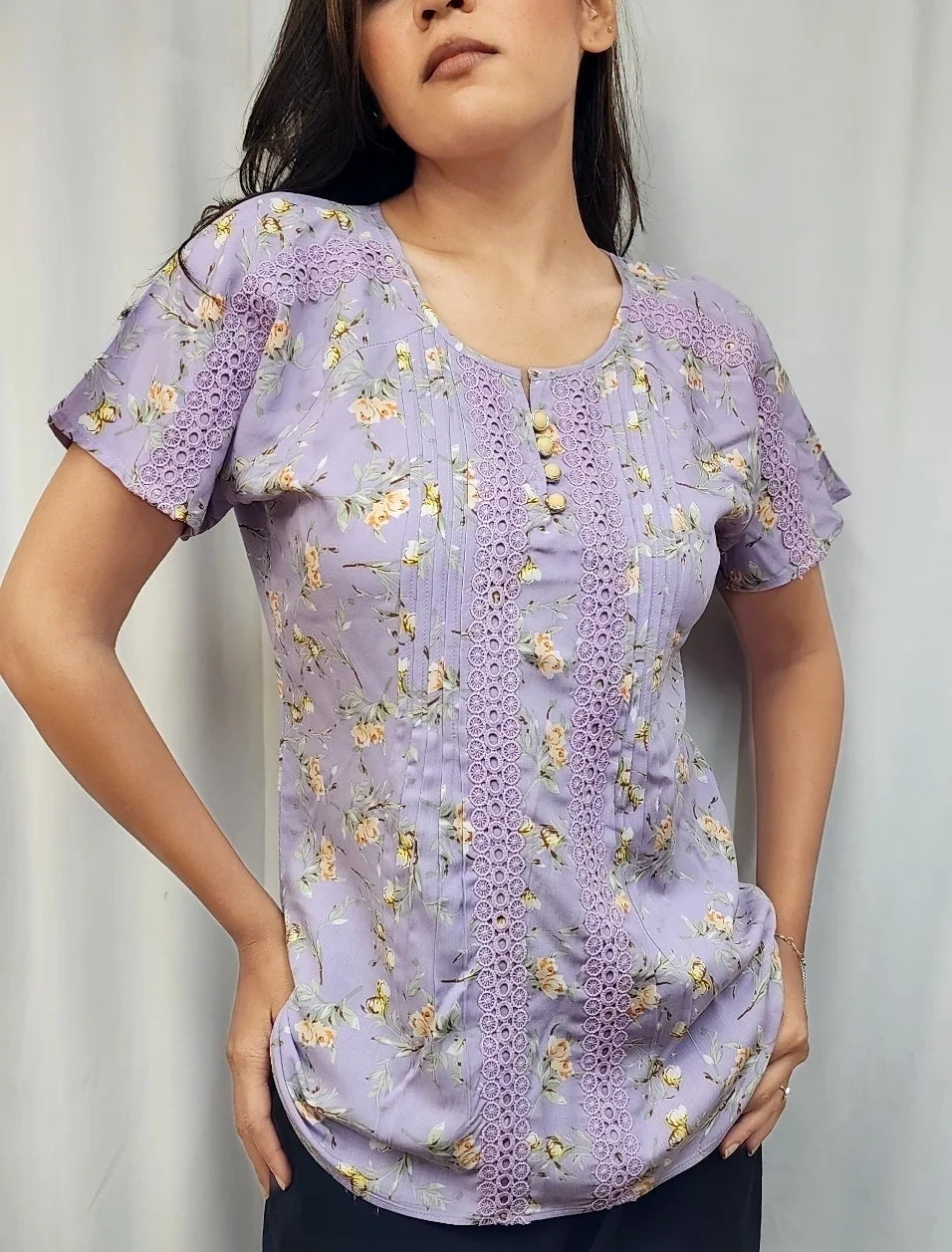 A woman modeling a light purple short-sleeve blouse with a floral print and a decorative embroidered front panel, paired with dark pants, set against a neutral light gray backdrop.