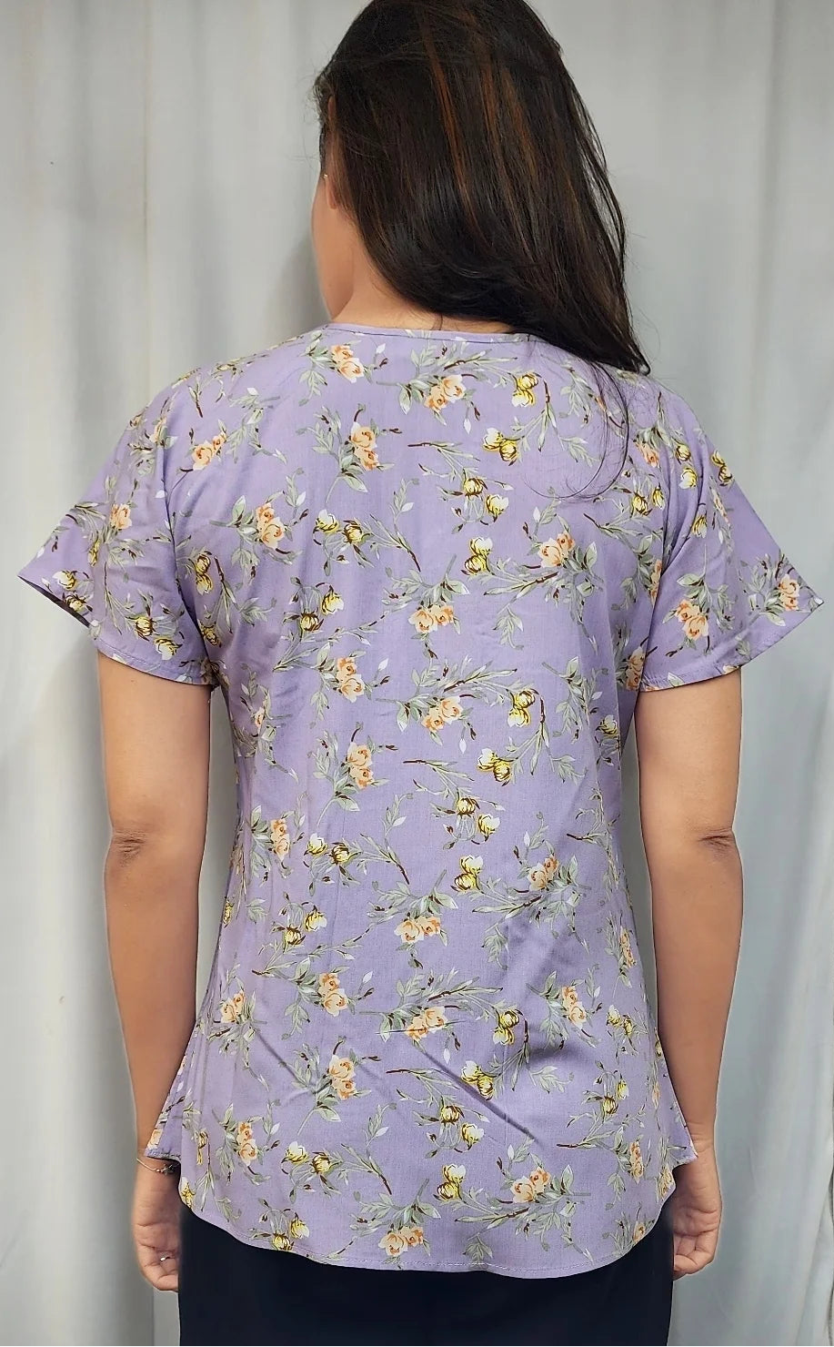 A woman viewed from behind wearing a lavender scrub top with a small yellow and white floral print, paired with black scrubs pants, set against a neutral curtain background.