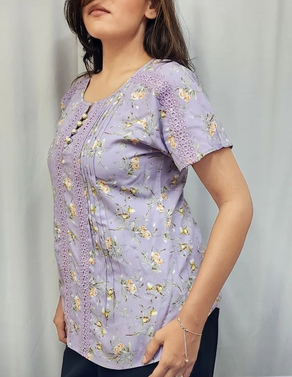 A woman modeling a light purple short-sleeve top with a yellow and green floral pattern, standing against a white curtain background.